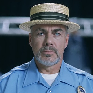 A man in a light blue police uniform with a straw boater hat and a stern expression looks directly at the camera. The…