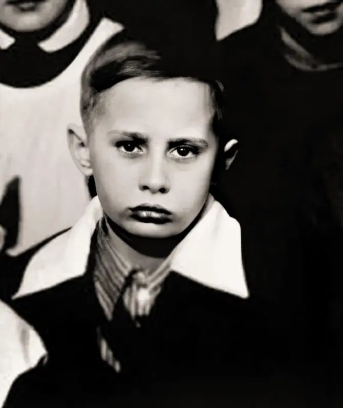 A black-and-white photograph of a young boy with a stern, unimpressed expression, wearing a collared shirt and tie, looking…