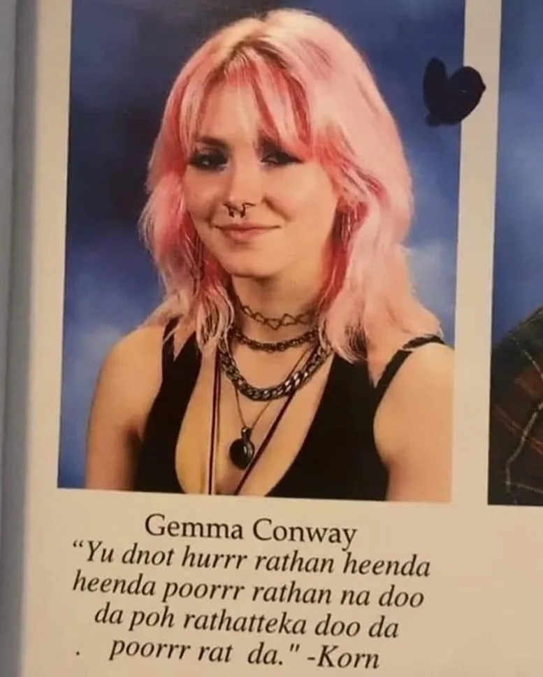 A school yearbook photo of a young woman with pink hair, a nose ring, and layered necklaces, smiling at the camera. Below…