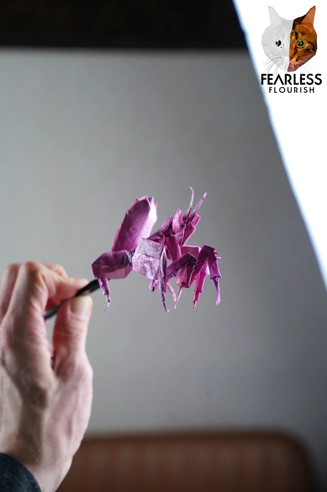 A hand holds a black tool, delicately positioning a complex, purple origami mantis. In the top right corner, a logo features…