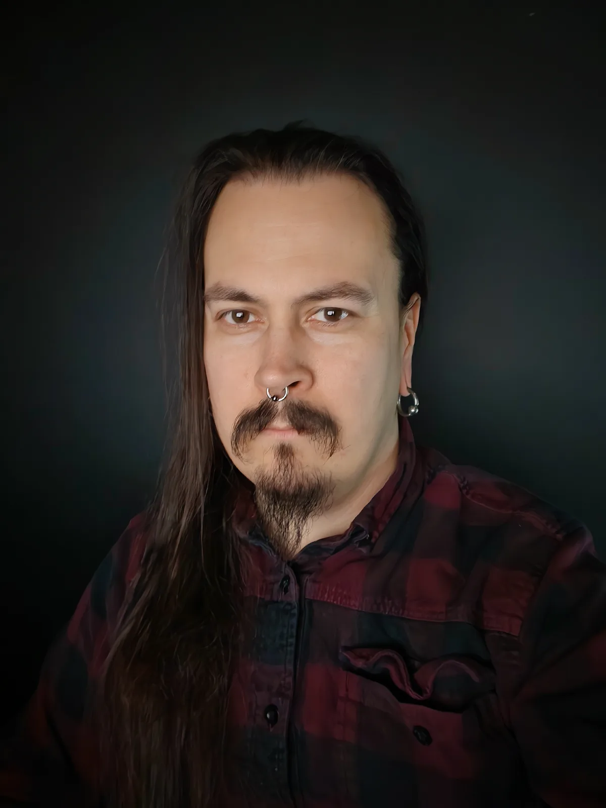 A man with long dark hair, a mustache, goatee, nose ring, and hoop earring stares directly at the camera with a serious…
