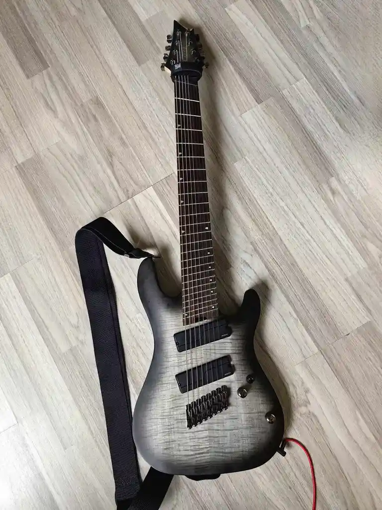 A black and grey electric guitar with a black strap lies on a light wood floor, with a red cable plugged into its jack. The…