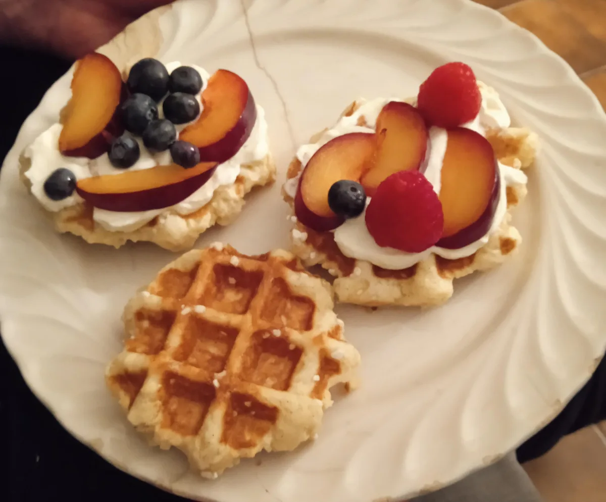 A white plate holds three small waffles; two are topped with whipped cream, sliced peaches, blueberries, and raspberries…