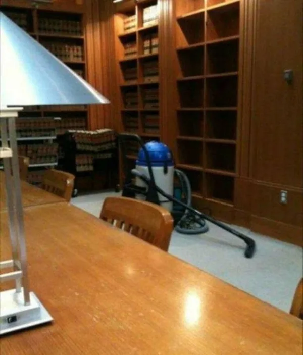 A blue and silver vacuum cleaner is sitting on a carpeted floor in a library, next to tall wooden bookshelves that are…