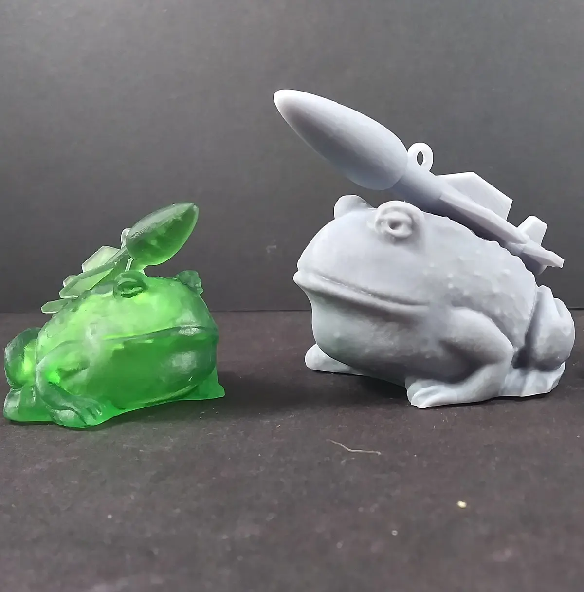 Two 3D-printed frog figurines sit side by side on a dark surface; the left is translucent green with a plant-like structure…