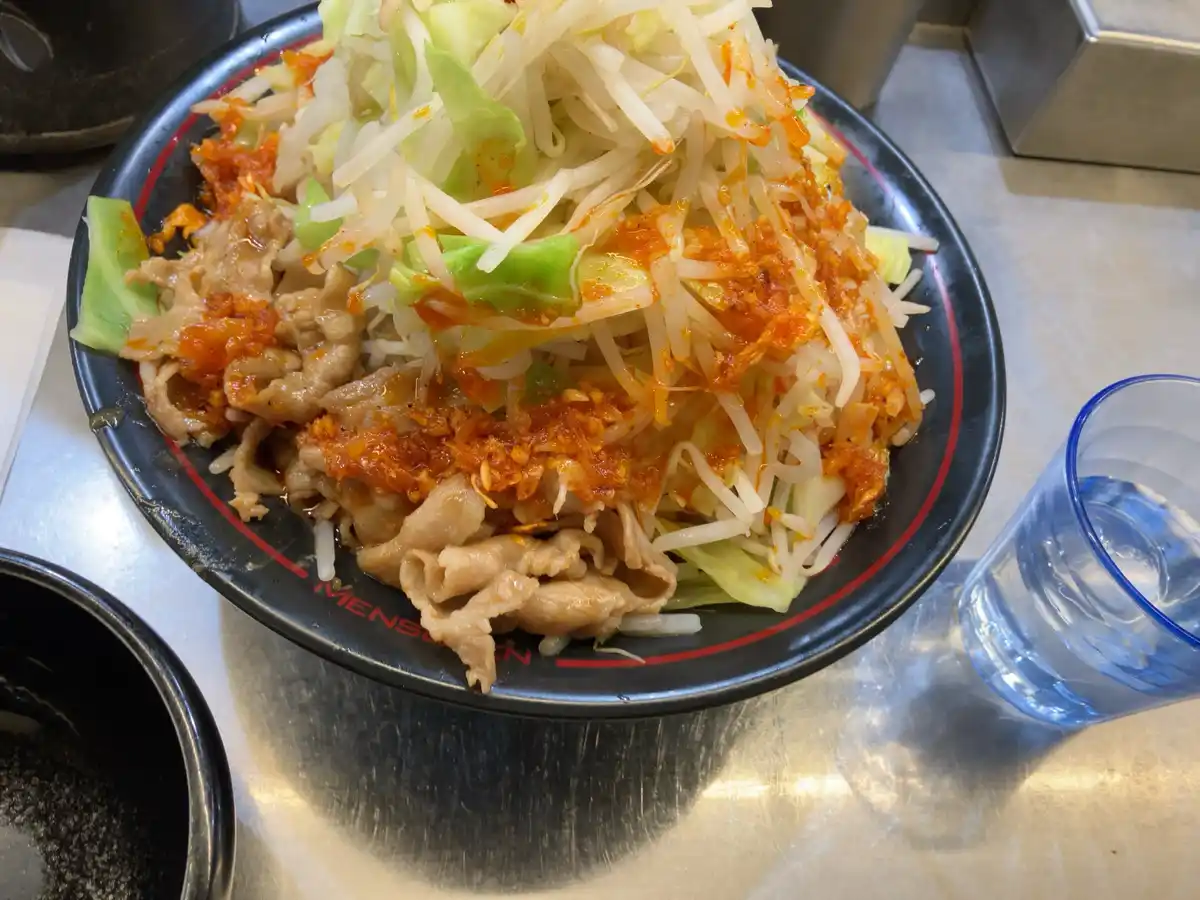 A close-up photo shows a black bowl with red trim, labeled 'MENSO', filled with sliced meat, shredded cabbage, and a large…
