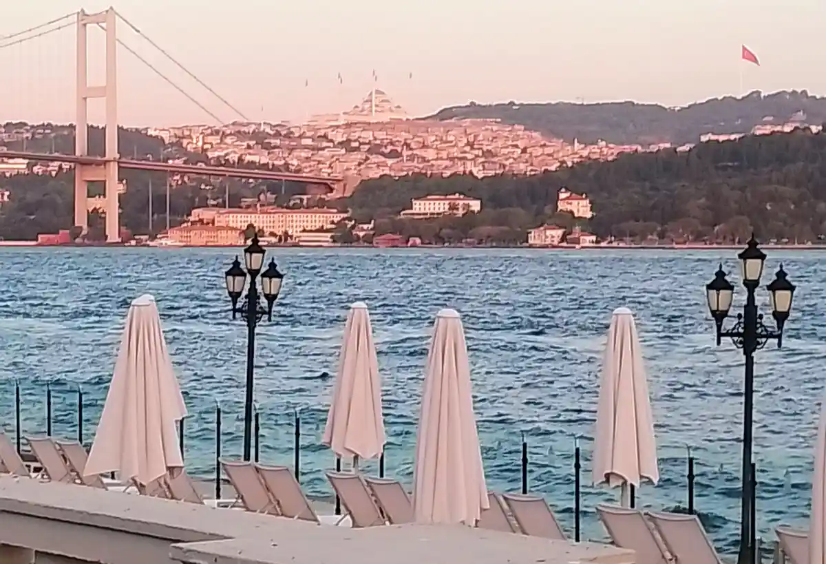 A scenic view of a waterfront with closed beige umbrellas and lounge chairs in the foreground, overlooking a body of water…