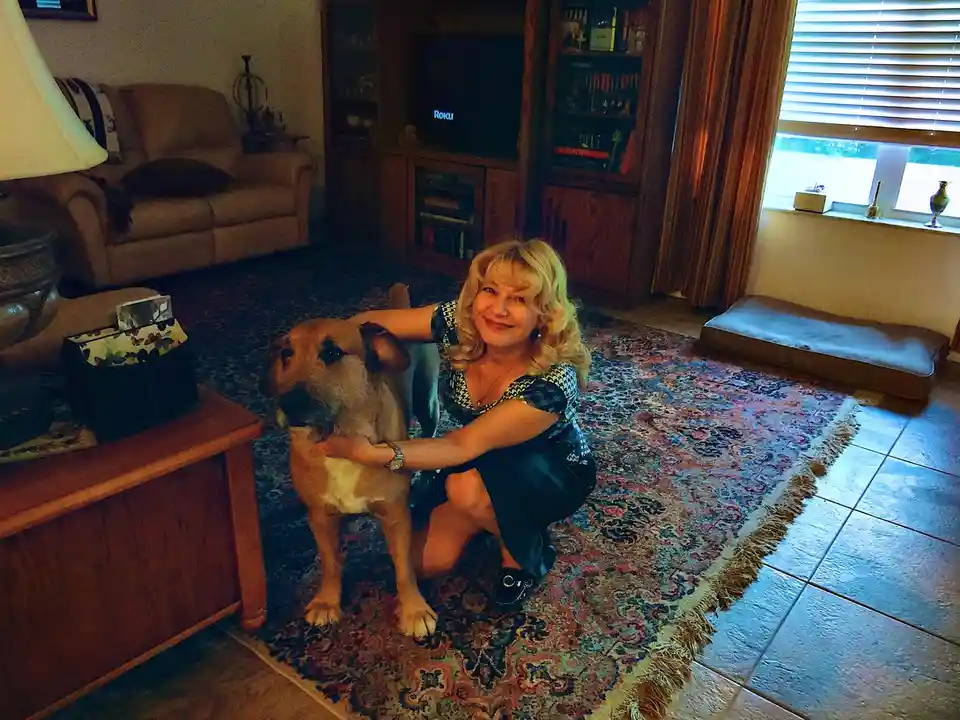 A woman with blonde hair is kneeling on a patterned rug in a living room, smiling at the camera while petting a medium-sized…