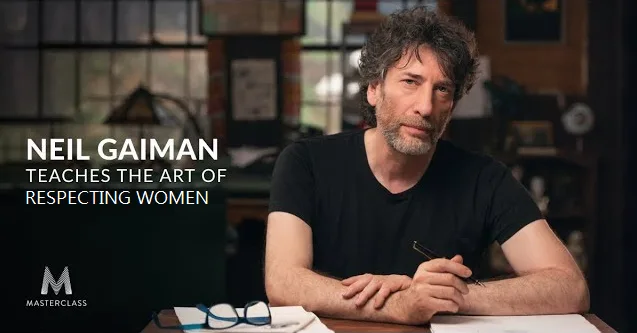 A man with curly dark hair and a beard, wearing a black t-shirt, sits at a wooden desk holding a pen. In the background…
