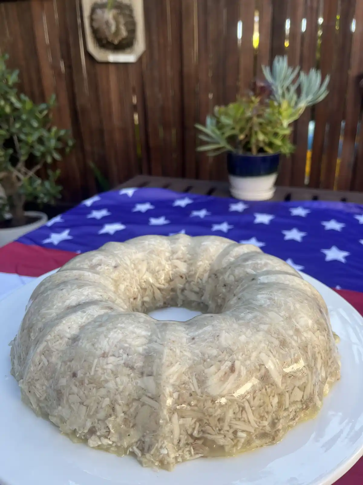 A bundt cake-shaped dessert covered in shredded coconut sits on a white plate atop an American flag spread on a table, with…