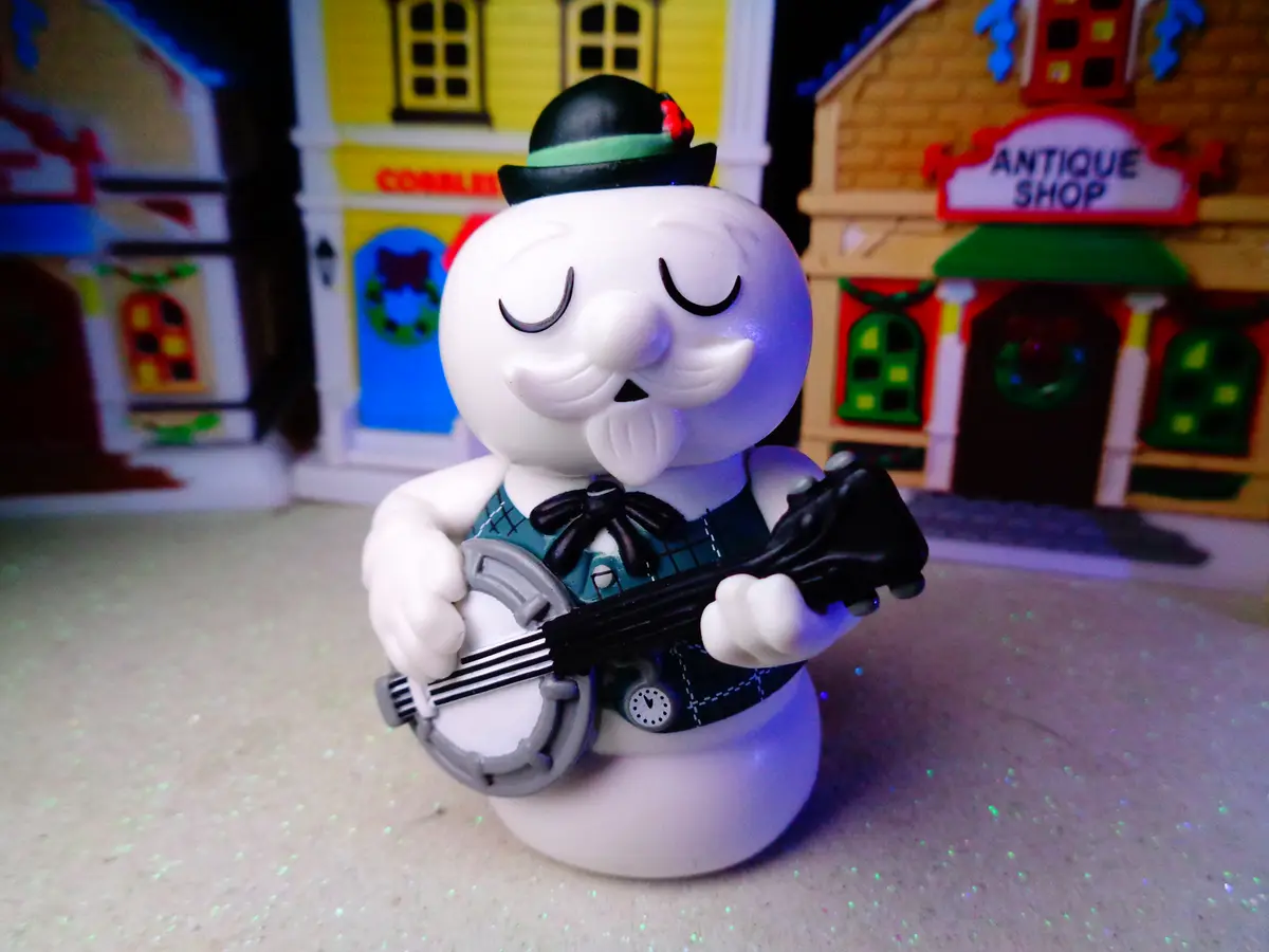 A white snowman figurine with a black top hat and green plaid vest is playing a banjo in front of miniature toy buildings…
