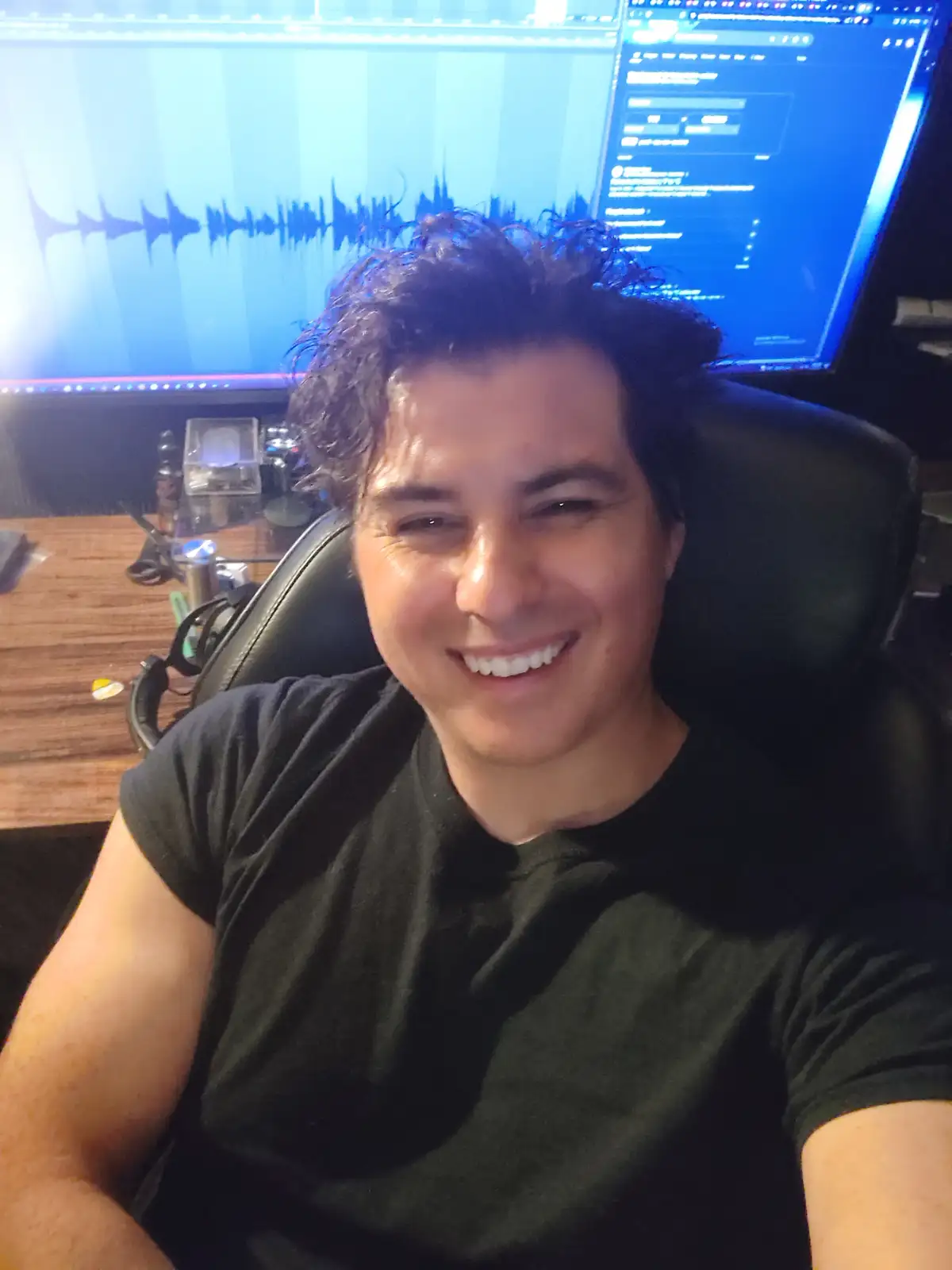 A man with dark curly hair and a wide smile sits in a black chair in front of a computer setup with two monitors. The left…