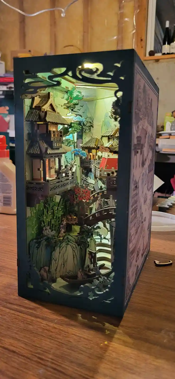 A detailed 3d paper craft diorama depicting a traditional chinese village scene with pagodas, bridges, boats, and miniature…