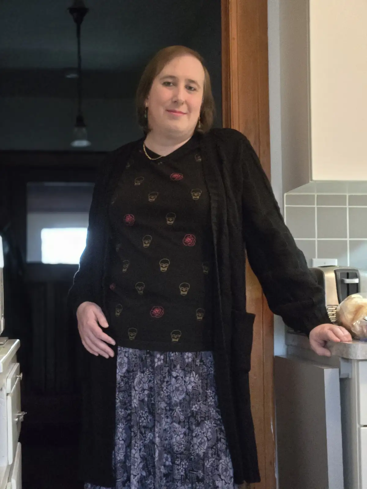 A woman stands in a kitchen, smiling slightly while leaning on a counter next to a toaster. She wears a black cardigan over…
