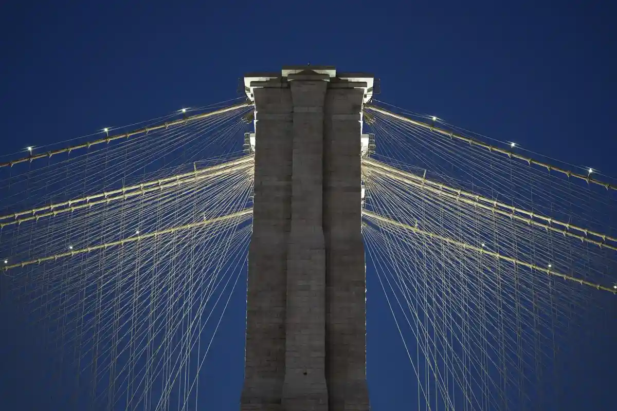 A low-angle night photograph of the Brooklyn Bridge's central stone tower, with its illuminated suspension cables radiating…