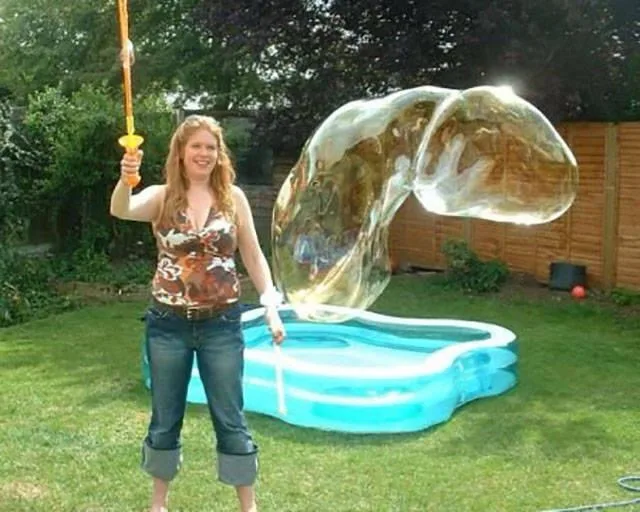 A woman stands in a grassy backyard next to a blue inflatable kiddie pool, holding a bubble wand. A massive, iridescent…
