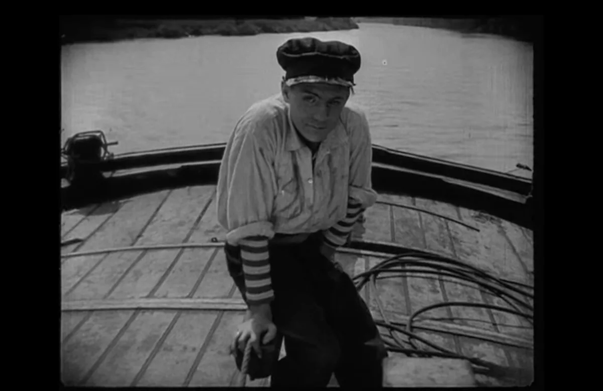 A black-and-white still shows a young man in a boat, wearing a cap and striped sleeves, looking directly at the camera with…