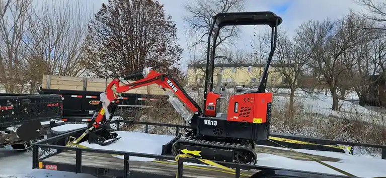 A small red and black mini excavator, model VA13, sits on a snowy trailer with yellow straps. The machine is partially…