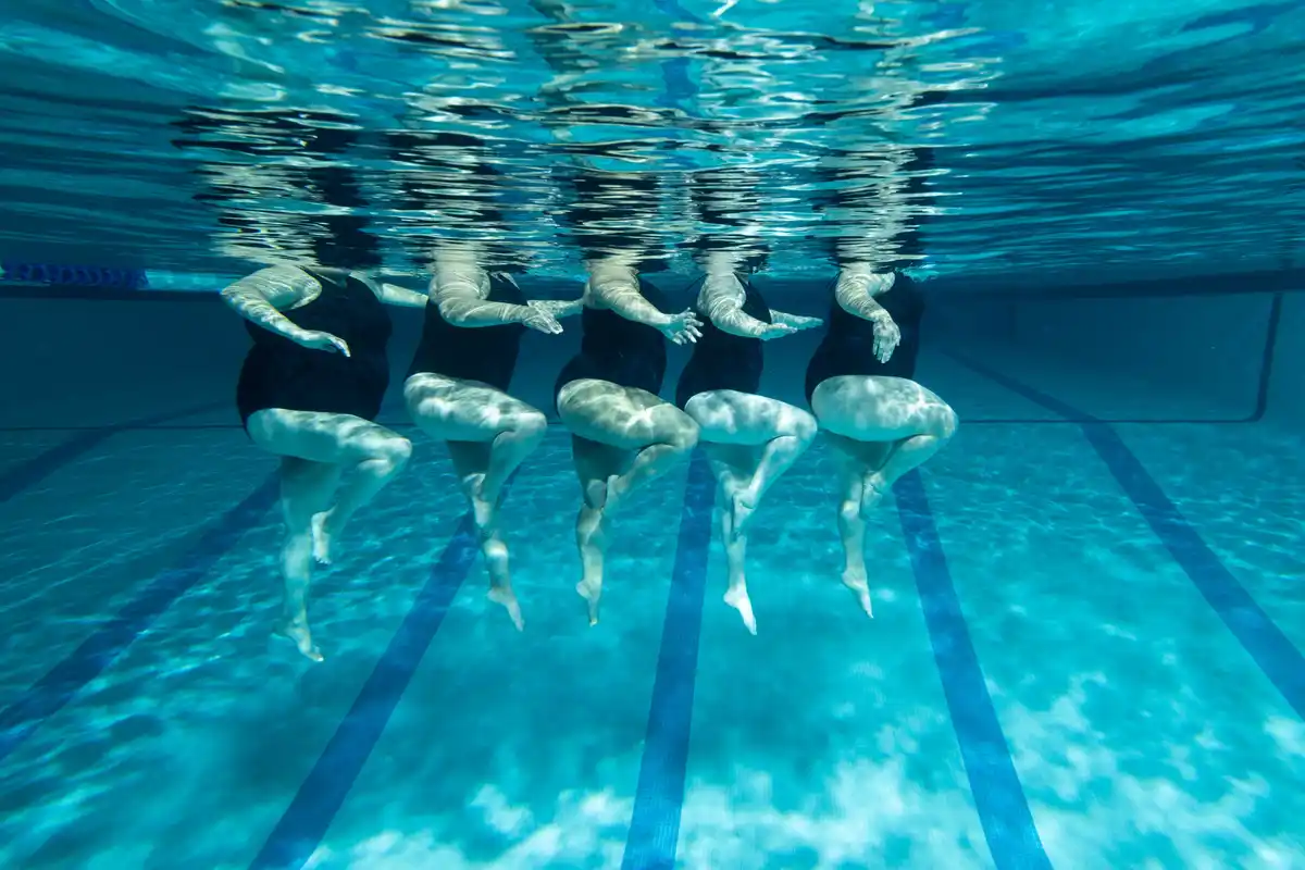 An underwater photo shows five people in black swimsuits floating in a pool lane, legs bent and arms extended, with sunlight…