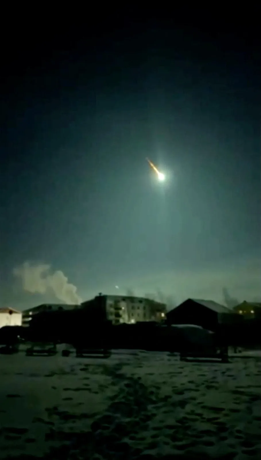 A blurry nighttime photo shows a bright, fiery object streaking across the sky above a snow-covered urban landscape with…