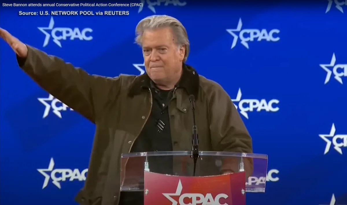 A man with gray hair wearing a brown jacket stands at a clear podium with a red CPAC logo, gesturing with his right arm…