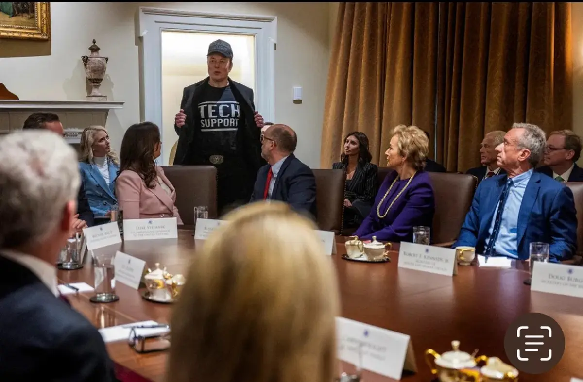 A man wearing a black t-shirt with 'TECH SUPPORT' printed in white letters stands in a formal meeting room, addressing…