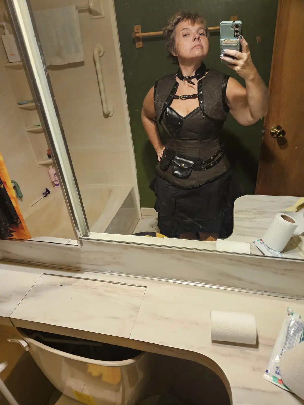 A woman in a dark, corseted, steampunk-style outfit takes a mirror selfie in a bathroom, holding her phone with one hand…