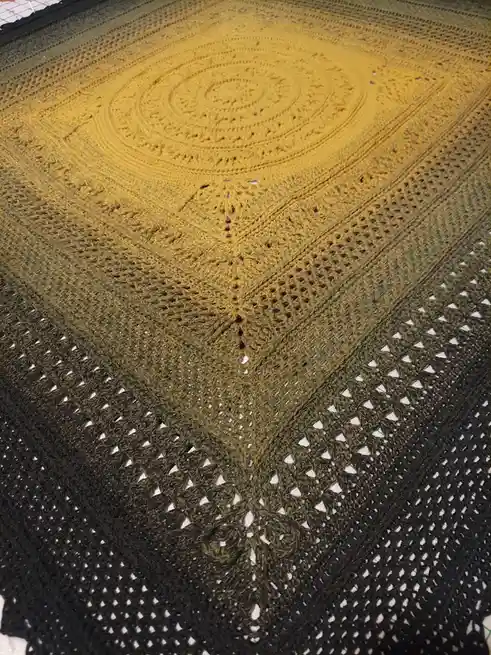 A close-up photo of a hand-crocheted shawl laid flat, featuring a gradient color transition from bright yellow at the center…