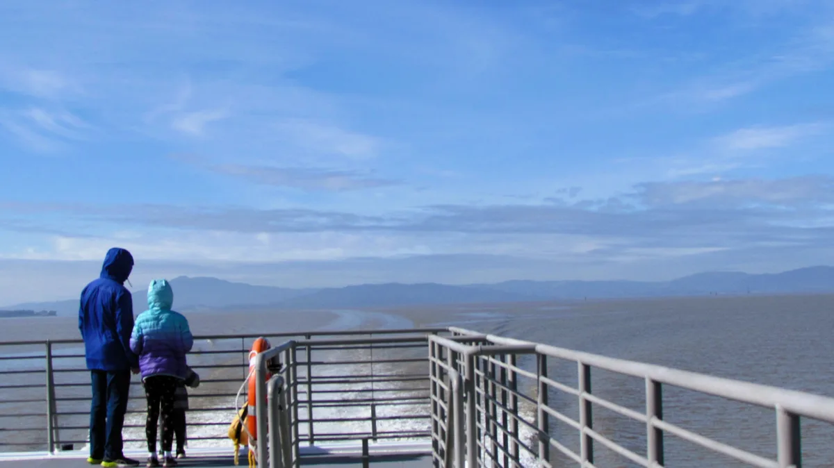 Two people, an adult and a child in hooded jackets, stand on a ferry deck overlooking a wide body of water with distant…