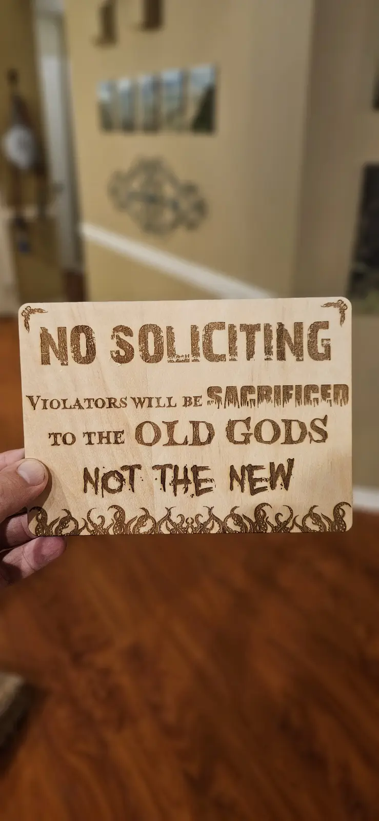 A hand holds a wooden sign with engraved text that reads 'NO SOLICITING. VIOLATORS WILL BE SACRIFICED TO THE OLD GODS NOT…