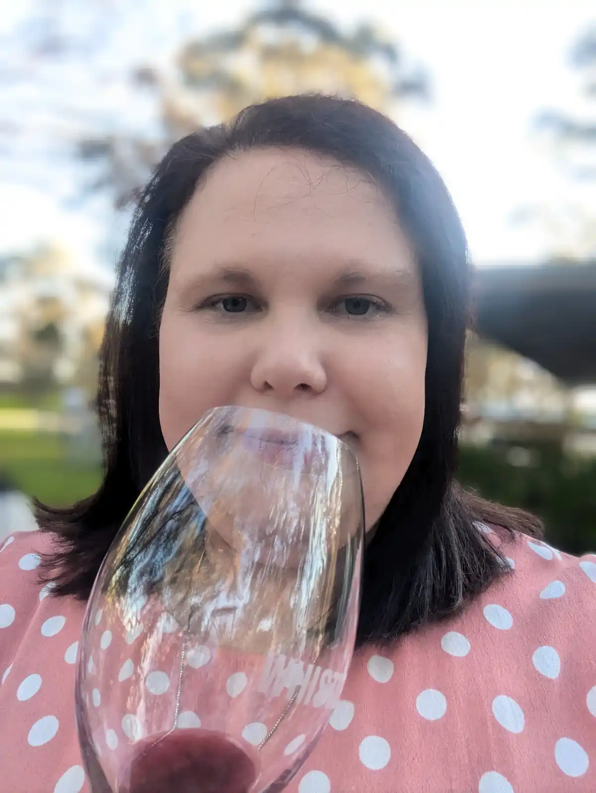 A woman with dark hair and a pink polka-dot shirt holds a wine glass to her lips, drinking red wine. The glass has a logo…