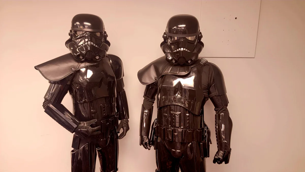 Two people in glossy black stormtrooper costumes stand side by side against a plain wall. The person on the left has a…