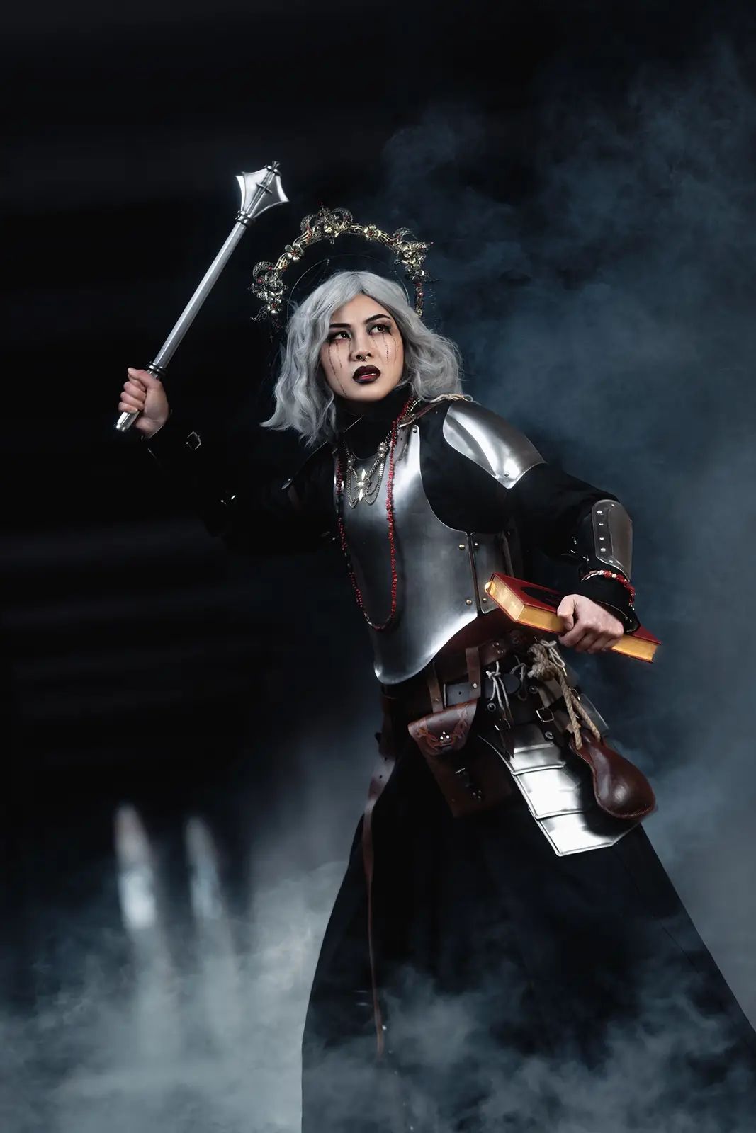 A person in dark fantasy cosplay, wearing silver armor and a crown, holds a hammer in one hand and a red book in the other…