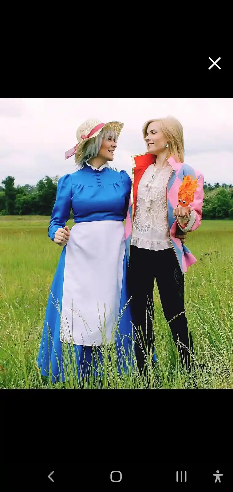 Two women in cosplay stand in a grassy field, one dressed as a maid in a blue dress and white apron with a straw hat, the…