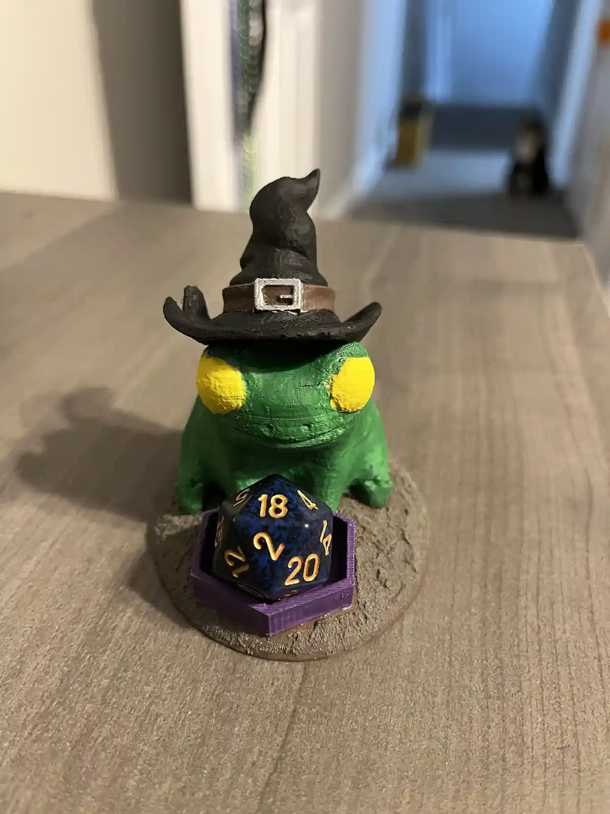 A green frog figurine wearing a black witch's hat with a silver buckle sits on a textured base. In front of it, a blue…