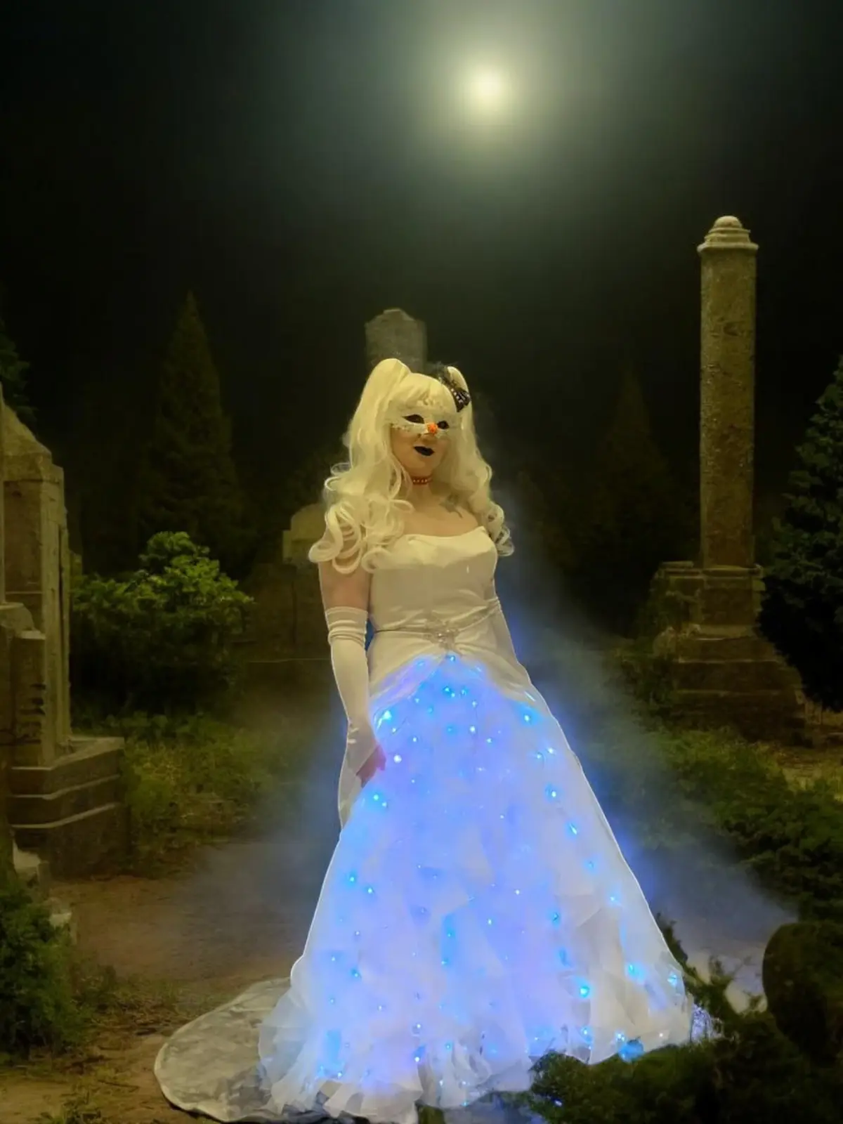 A person in a white wedding gown with glowing blue lights on the skirt stands in a foggy graveyard at night, wearing a white…