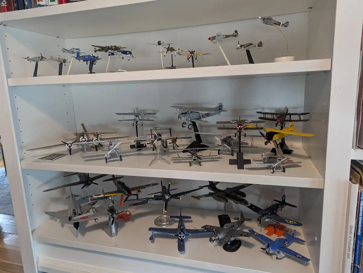 A white bookshelf with three shelves displays numerous detailed model airplanes of various types and colors, including…