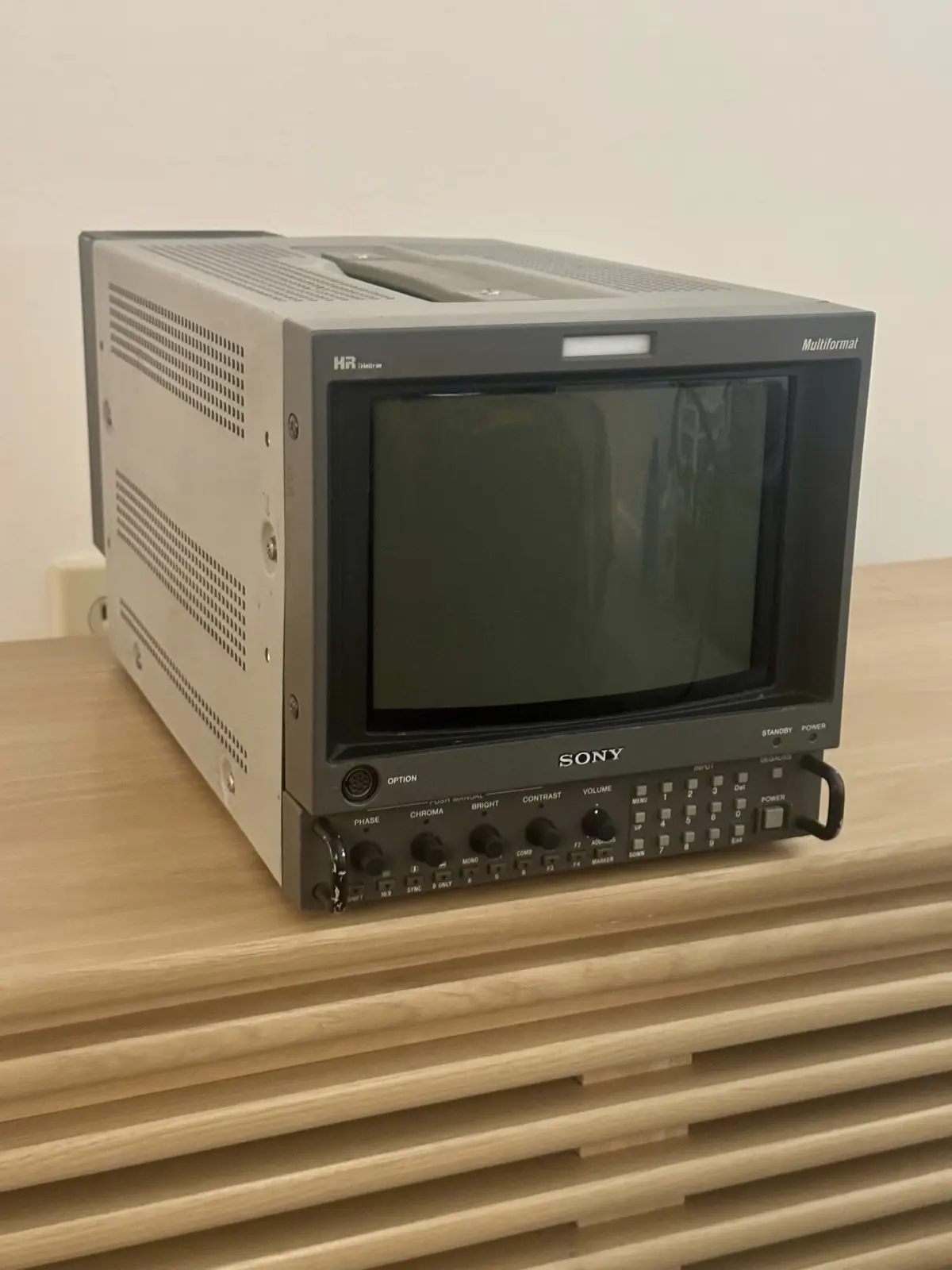 A vintage Sony HR Multiformat video monitor sits on a modern wooden slatted shelf, its screen dark and unpowered, with…