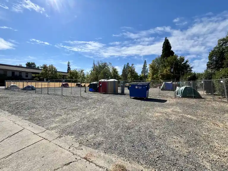A gravel lot enclosed by a chain-link fence contains several tents, portable toilets, and a blue dumpster under a bright…