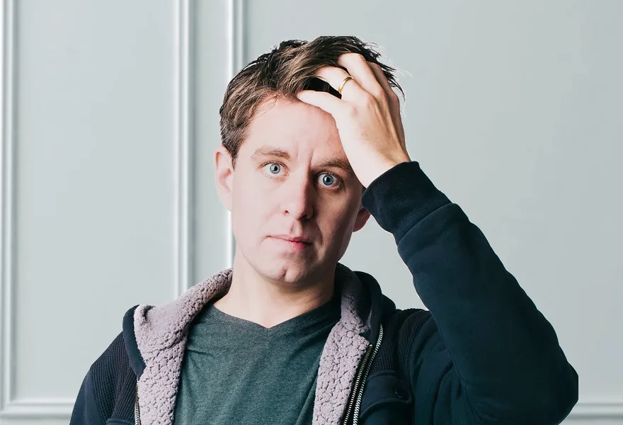 a man with blue eyes and brown hair, wearing a dark hoodie with a light-colored fleece-lined hood, looks directly at the…