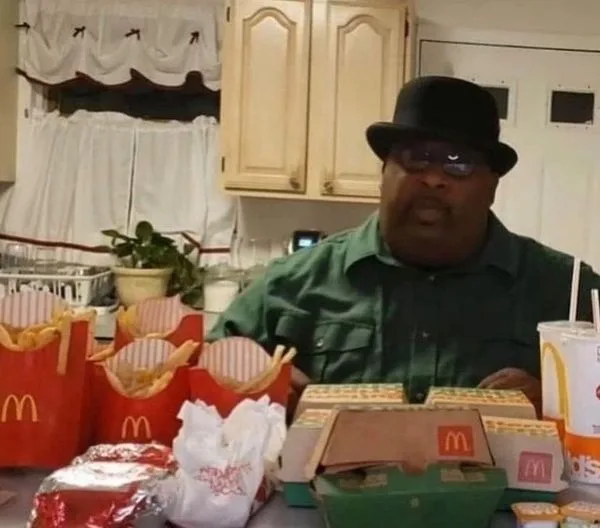 a man wearing a black fedora and glasses sits at a kitchen table surrounded by numerous McDonald's food items including…