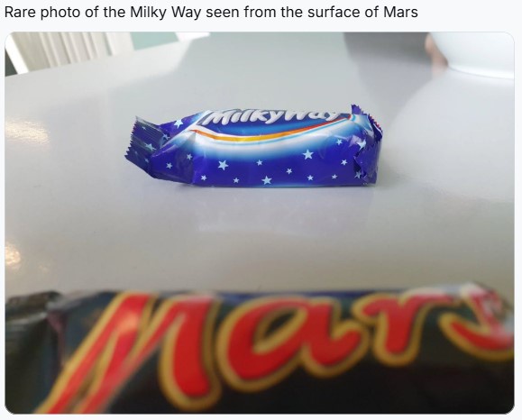 A close-up photo shows a blue-wrapped Milky Way chocolate bar on a white surface, with a blurred Mars chocolate bar in the…