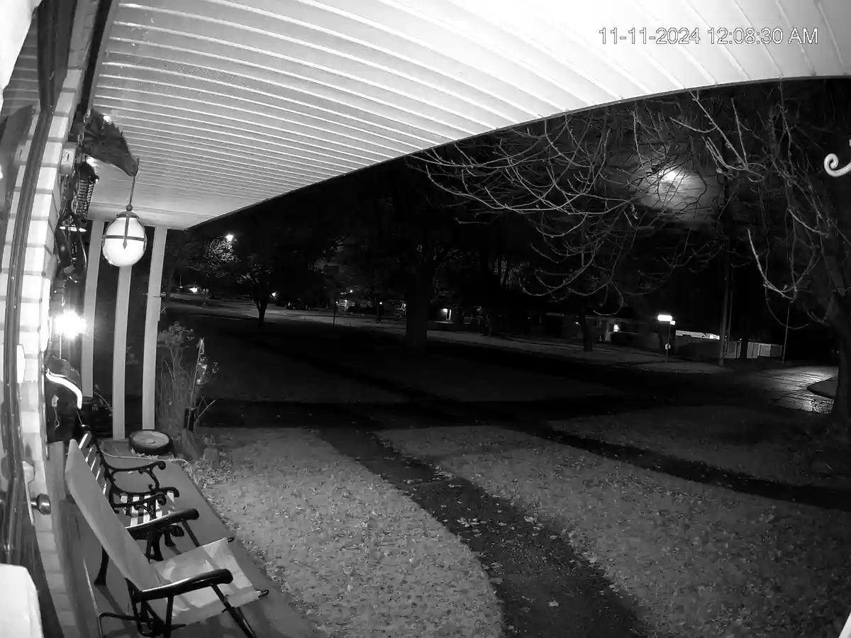A black-and-white nighttime security camera photo from a porch, showing a quiet street with bare trees, a lit porch light…