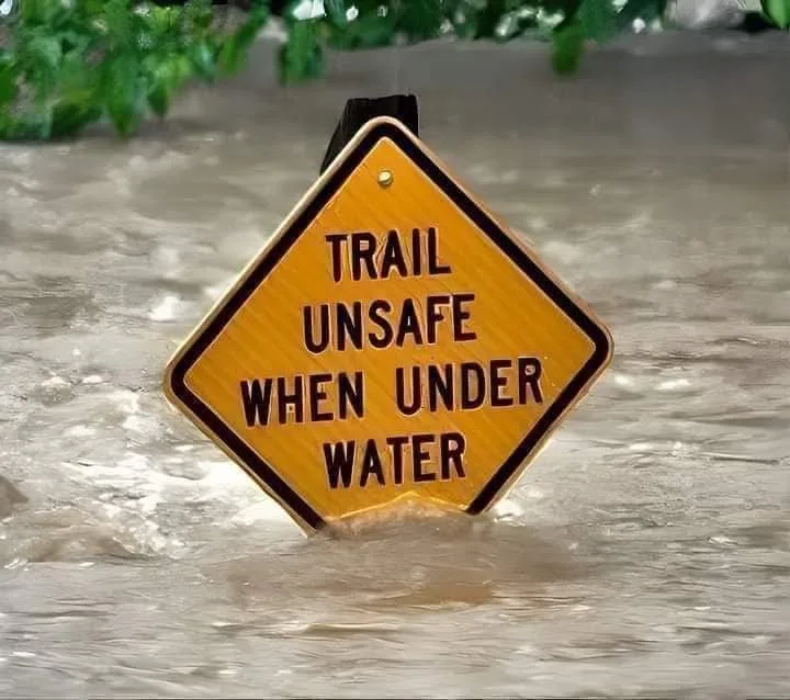 A yellow diamond-shaped warning sign with black text reading 'TRAIL UNSAFE WHEN UNDER WATER' is partially submerged in murky…