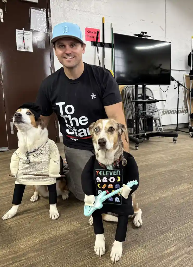 A man in a blue cap and black t-shirt stands behind two dogs dressed in human-like costumes. The dog on the left wears a…