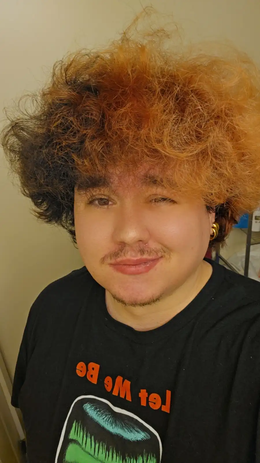 A person with a large, curly, two-toned hairstyle—dark brown on the sides and bright orange on top—is smiling slightly while…