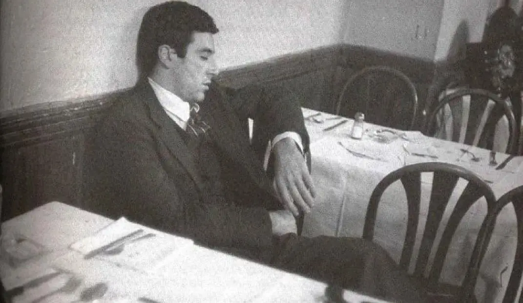 A black-and-white photo shows a man in a suit slumped over at a restaurant table, appearing asleep or extremely bored. The…