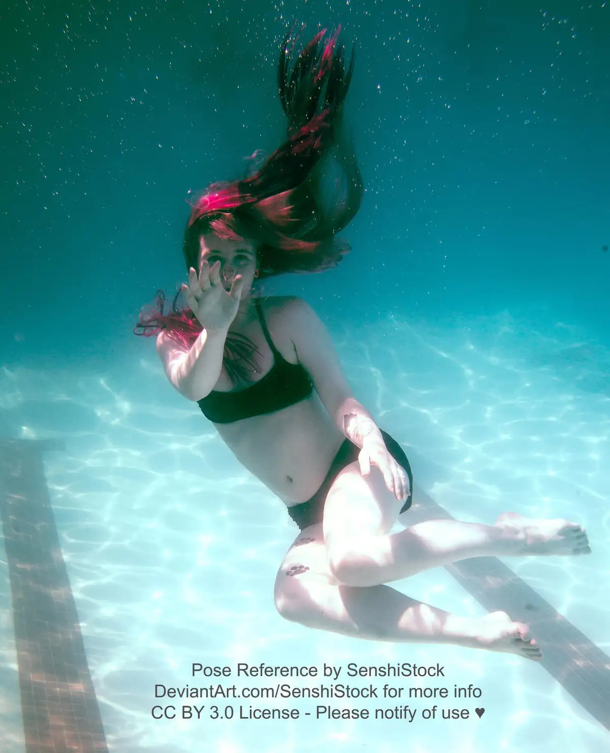 An underwater photograph shows a woman with long dark hair streaked with pink, wearing a black bikini, floating in a pool…
