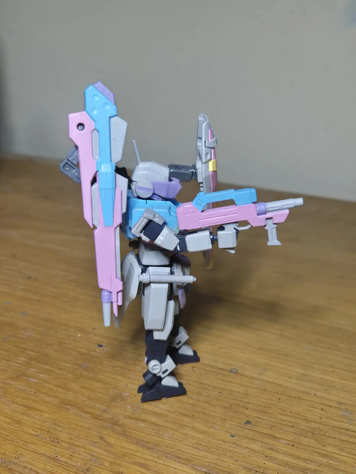 A plastic robot model, resembling a Gundam, stands on a wooden surface with its body painted in gray, blue, and purple…