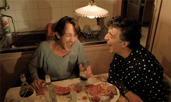 A six-panel image sequence shows two men at a kitchen table, initially laughing hysterically, then one man leans over to…